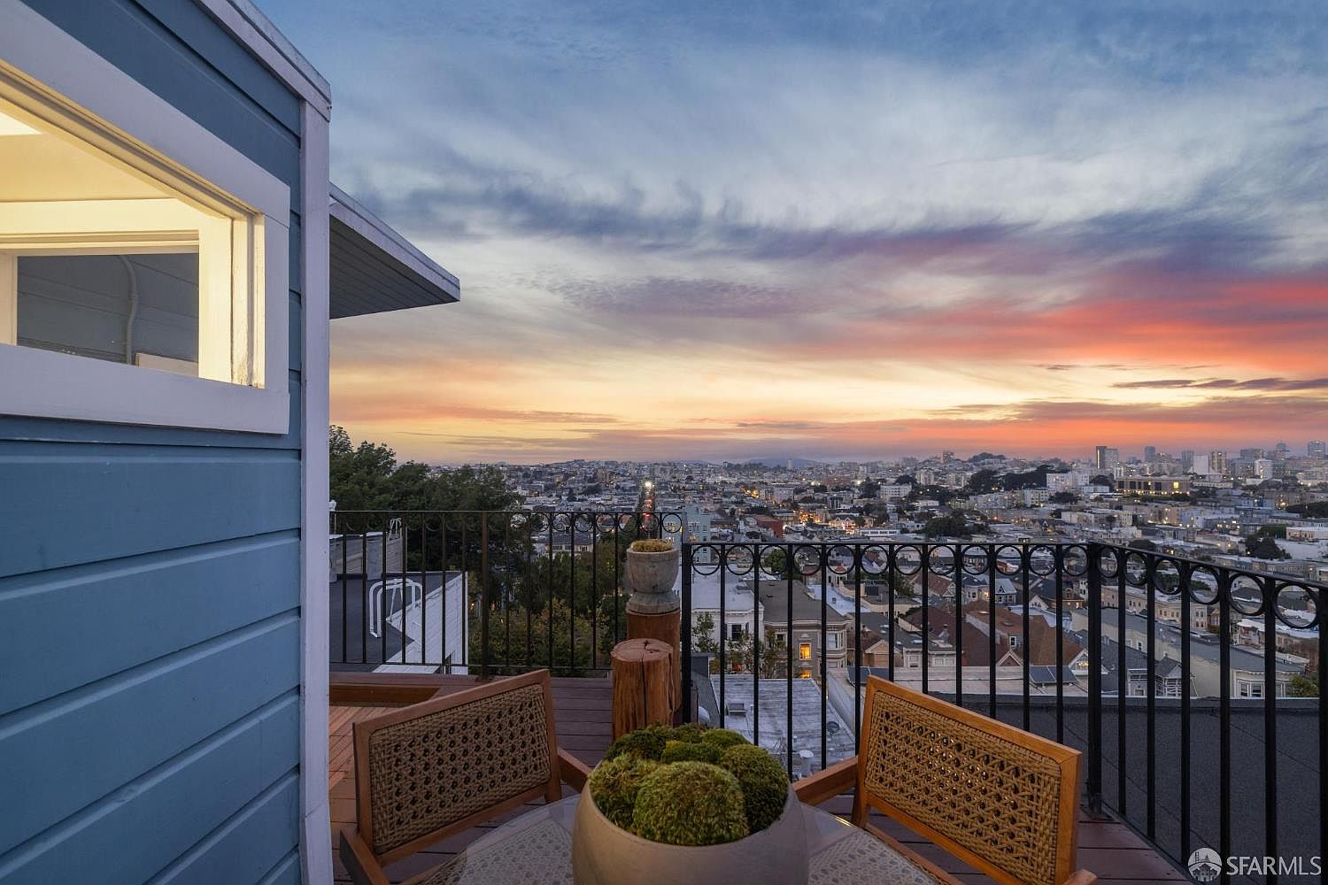 This image showcases a charming outdoor balcony or deck area with a stunning cityscape view at sunset. The space is furnished with wicker chairs and a small table, creating an inviting atmosphere for relaxation. The wrought iron railing adds a touch of elegance, while the vibrant sky enhances the overall appeal of this outdoor living space.