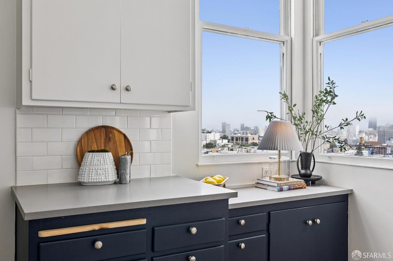 This is a well-lit kitchen corner featuring navy blue lower cabinets with silver knobs and a light gray countertop. White subway tile backsplash complements the white upper cabinets. A large window provides a view of the city skyline, enhancing the kitchen's appeal.