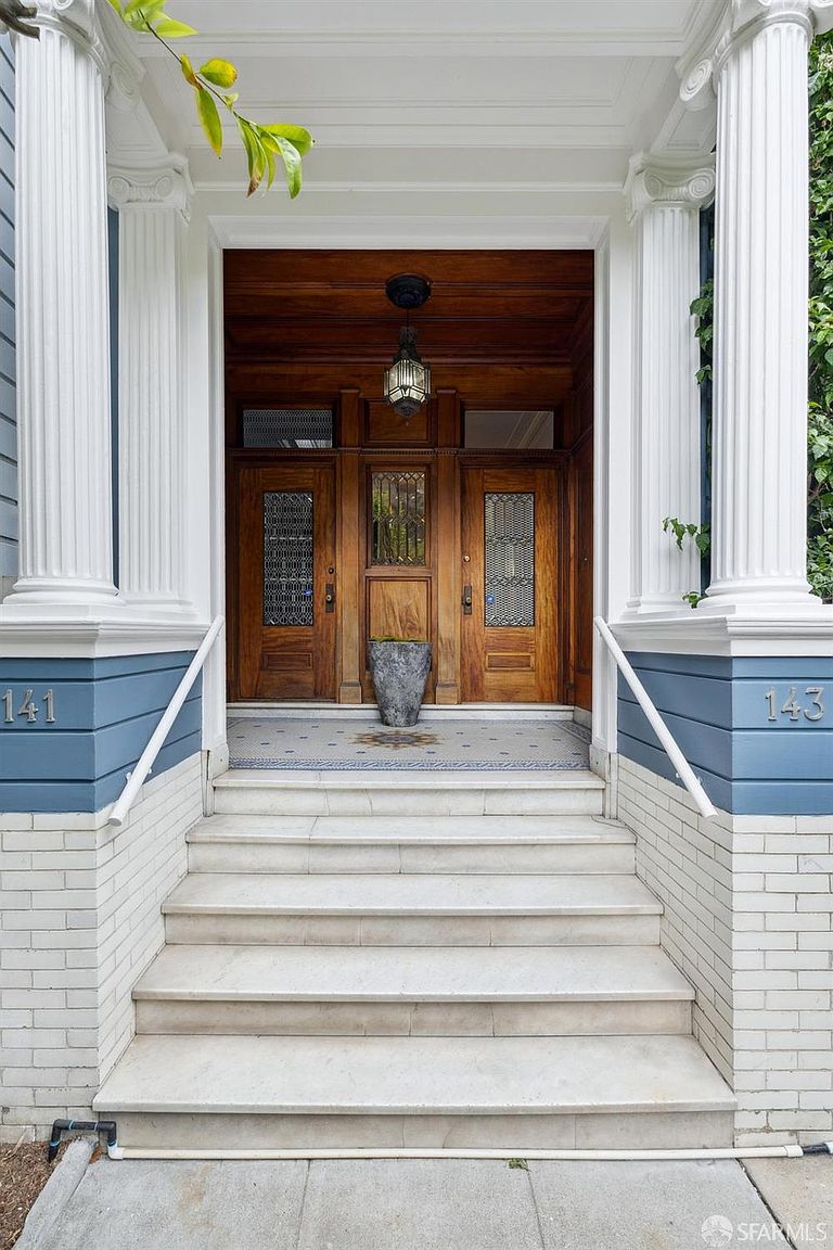 This is a welcoming entryway to a building, featuring a grand staircase leading up to three wooden doors. The entrance is framed by white columns and a blue facade with the numbers 141 and 143 displayed on either side. A decorative light fixture hangs above the doors, adding to the classic and elegant aesthetic.