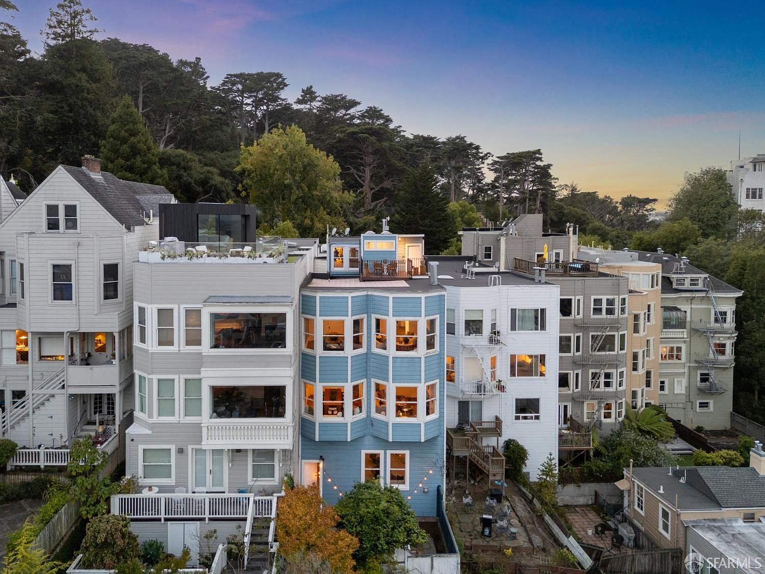 This aerial view showcases a row of multi-story townhouses with varying architectural styles and colors, set against a backdrop of lush greenery and a twilight sky. The blue townhouse in the center stands out with its bay windows and string lights, while the surrounding buildings feature rooftop decks and fire escapes, creating a vibrant urban scene. The image provides a comprehensive view of the properties and their surroundings.
