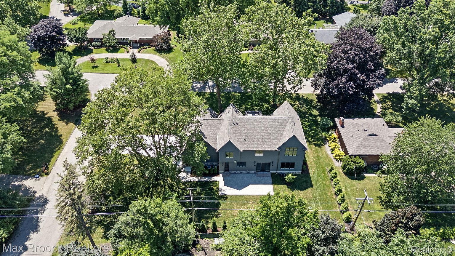 This aerial shot showcases a modern home with a gray exterior and a well-maintained lawn. The house features a unique roof design with multiple gables and a spacious patio area. Mature trees surround the property, providing shade and privacy, while neighboring houses are visible in the background, indicating a residential neighborhood.