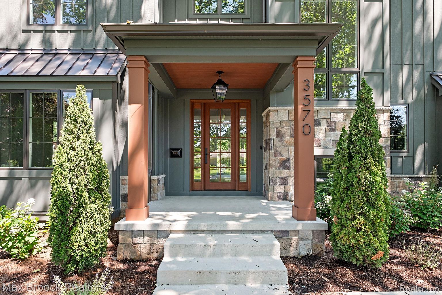 This is a well-lit, inviting entryway to a home. The entrance features a double door with glass panes, flanked by stone-clad columns and manicured evergreen trees. The house number '3570' is prominently displayed on one of the columns, adding to the curb appeal.