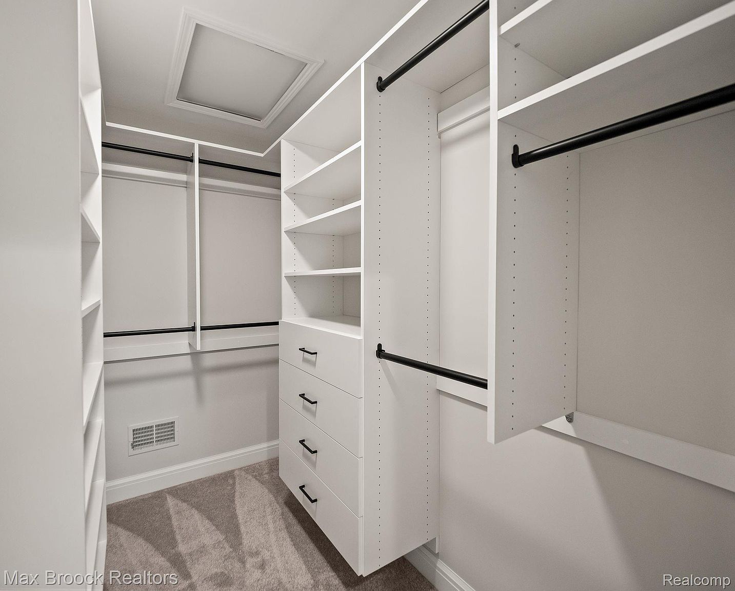 This is a well-organized walk-in closet featuring white shelving units, drawers with black hardware, and black closet rods. The closet is carpeted and has a clean, modern aesthetic. An access panel is visible in the ceiling.