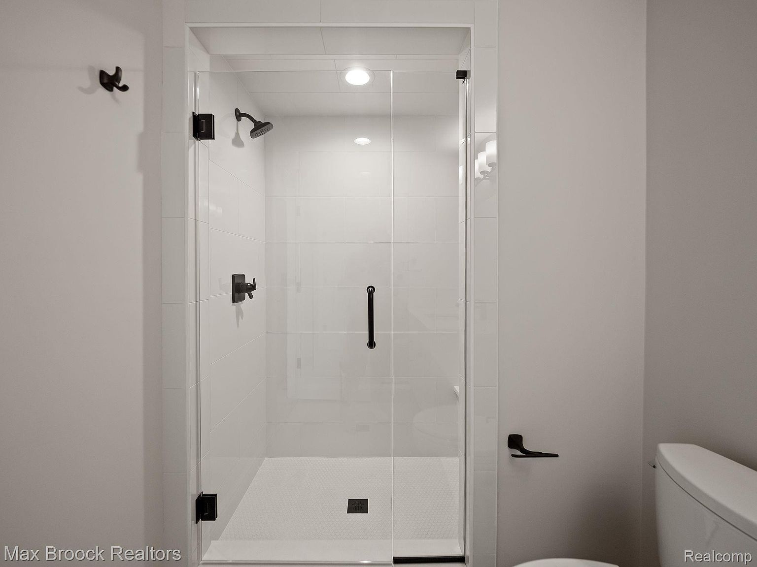 This is a well-lit bathroom featuring a modern glass-enclosed shower with white tile walls and a contrasting dark hardware. The shower has a sleek design with a rainfall showerhead and a dark handle on the glass door. The bathroom has a clean and minimalist aesthetic.