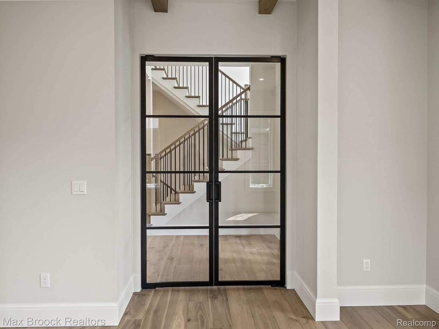 The image showcases an interior hallway featuring a staircase visible through a set of modern, black-framed glass doors. The staircase has wooden steps and black metal railings, creating a contemporary aesthetic. The walls are painted in a neutral tone, and the flooring is light-colored hardwood, contributing to a bright and airy atmosphere.