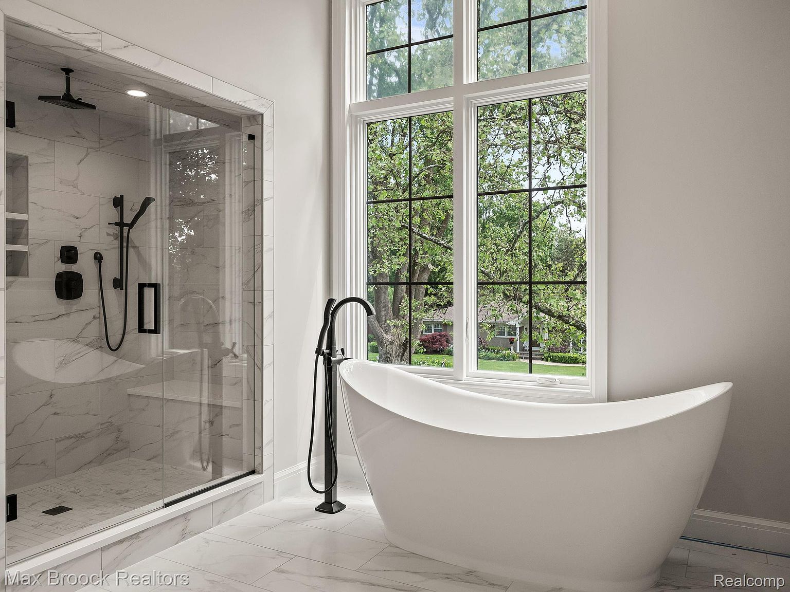 This is a luxurious primary bathroom featuring a freestanding white bathtub positioned in front of a large window with a view of the outdoors. A modern, black faucet stands next to the tub. The shower is enclosed in glass with black hardware and marble-like tile, creating a spa-like atmosphere.