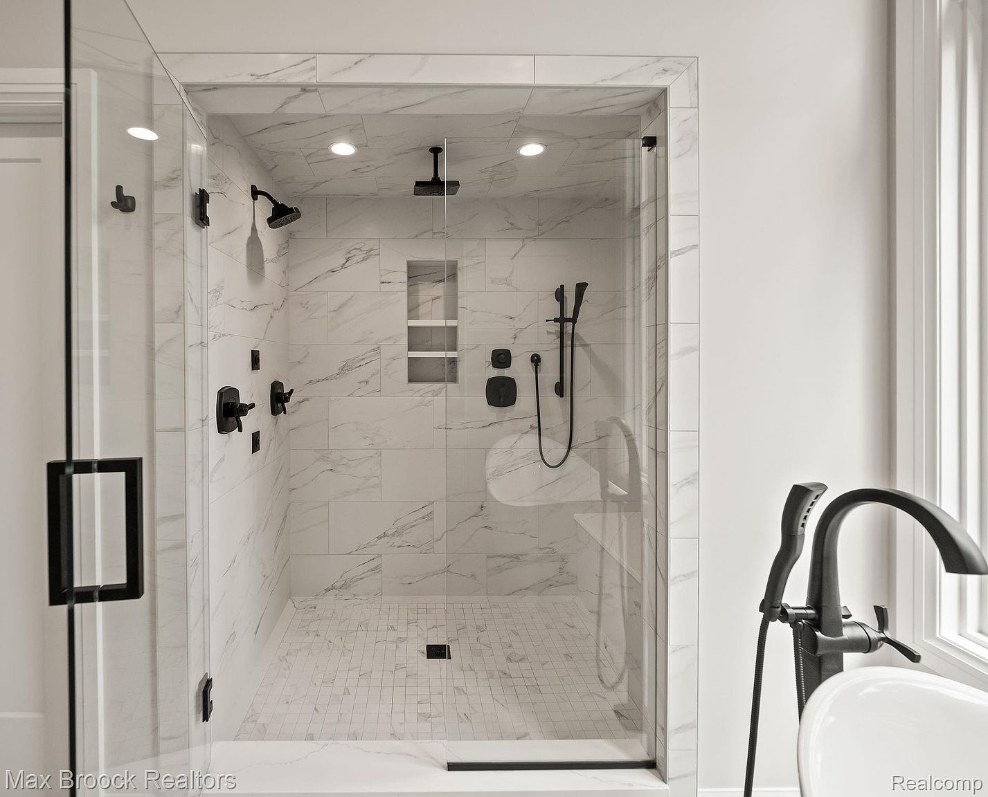 This is a well-lit primary bathroom showcasing a modern shower with marble-like tile walls and a mosaic tile floor. The shower features black fixtures, including a rainfall showerhead and handheld sprayer, along with a built-in niche for storage. The glass enclosure adds to the sleek and contemporary design.
