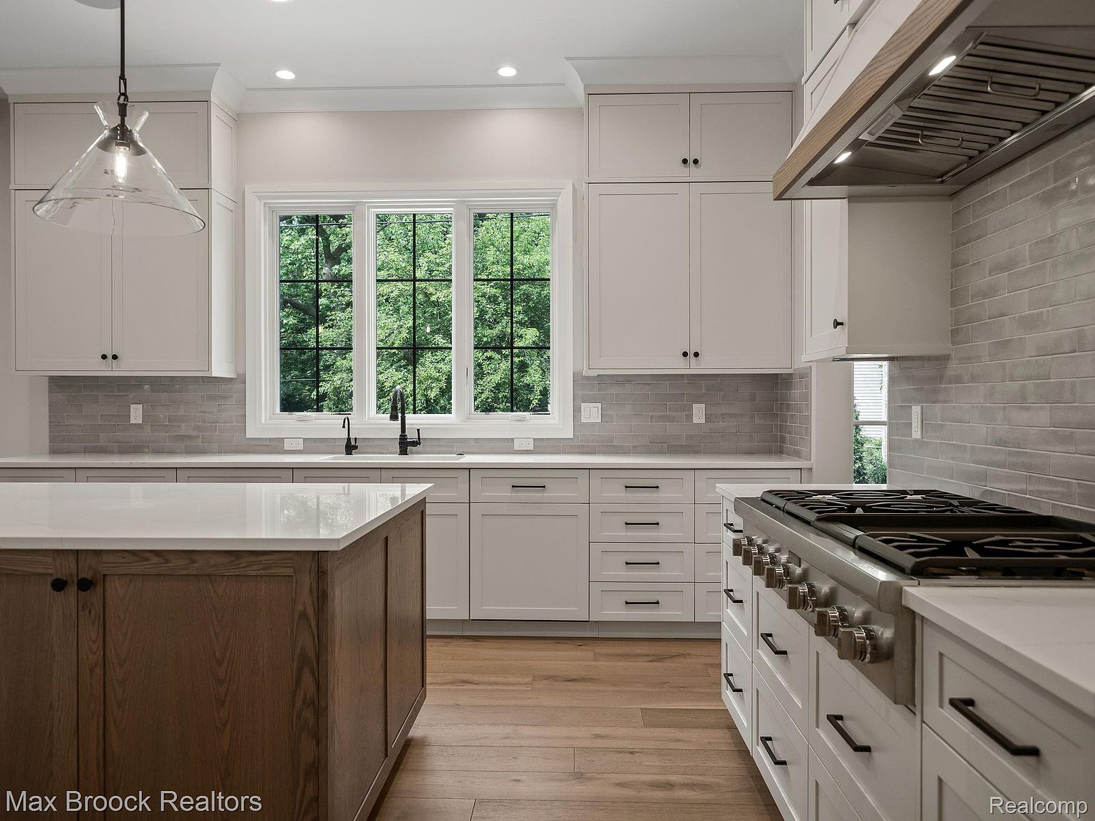 This is a well-lit kitchen featuring white cabinets, a large island with a wooden base and white countertop, and stainless steel appliances. A large window provides natural light and a view of the outdoors. The kitchen has a modern and clean design with a focus on functionality and style, as seen from a medium shot perspective.