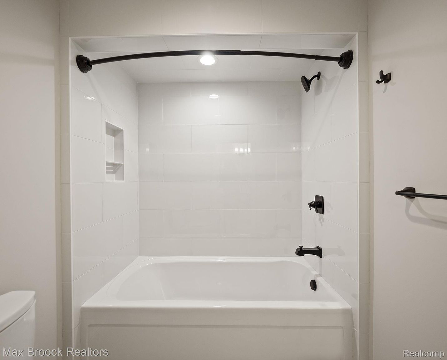 This is a well-lit bathroom featuring a white bathtub with a black faucet and shower head. The walls are tiled in white, and there is a built-in niche for storage. The bathroom also includes a black towel rack and a curved shower curtain rod, creating a clean and modern aesthetic.