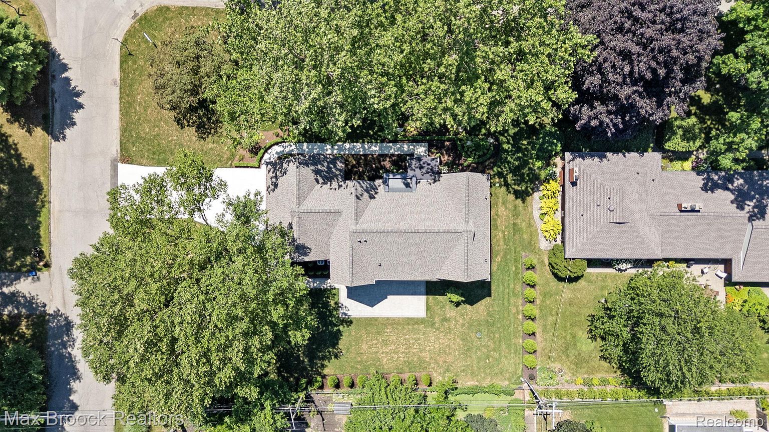 This aerial shot showcases a well-maintained single-family home with a gray roof, surrounded by lush greenery and mature trees. A concrete patio extends from the back of the house, leading to a manicured lawn. The property includes a driveway and is situated in a quiet, residential neighborhood.