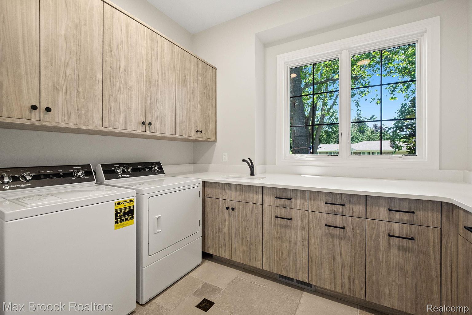 This is a well-organized laundry room featuring light wood cabinetry above and below a white countertop. A modern black faucet is installed in the sink. A front-loading washer and dryer are present, and a window provides natural light, enhancing the room's functionality and aesthetic appeal.