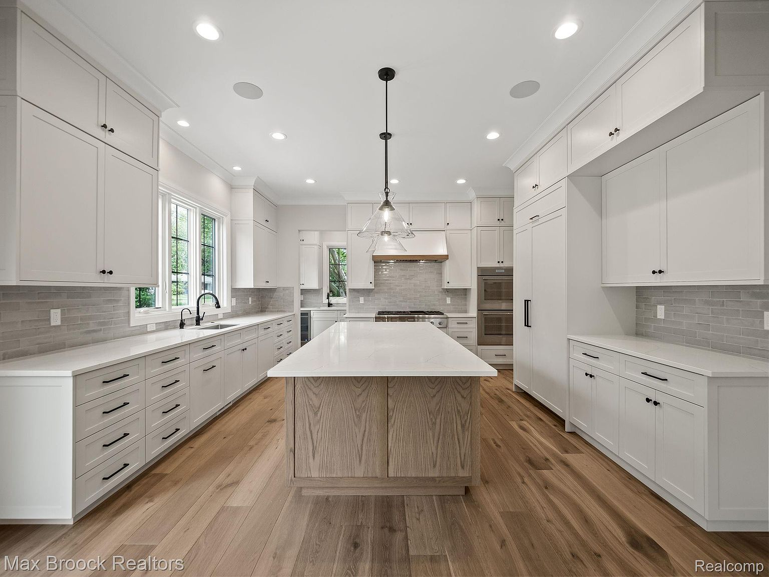 This is a bright and spacious kitchen featuring white cabinetry, a large center island with a light wood base and white countertop, and stainless steel appliances. The kitchen is well-lit with recessed lighting and pendant lights over the island, complemented by hardwood flooring. The design offers a clean and modern aesthetic, perfect for cooking and entertaining.