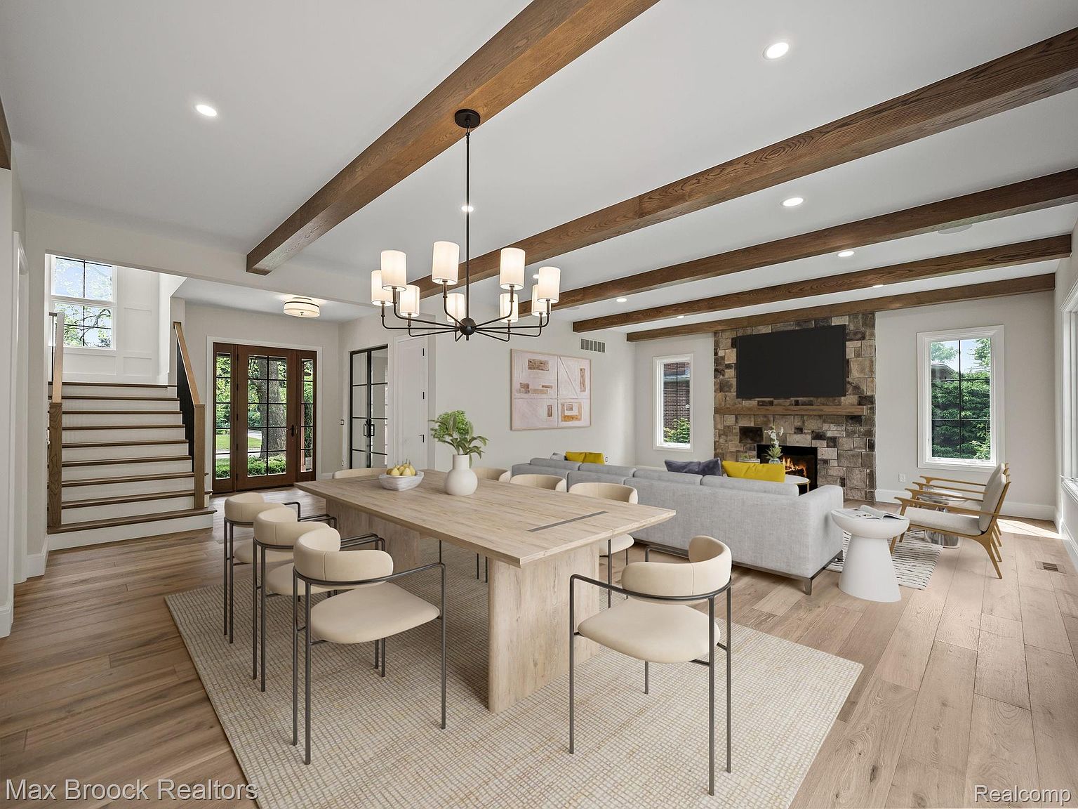 This interior shot showcases a dining room seamlessly connected to a living area. The dining space features a large wooden table surrounded by modern chairs, illuminated by a stylish chandelier. The living area includes a stone fireplace with a mounted TV, a comfortable sofa, and natural light streaming through the windows, creating a warm and inviting atmosphere.