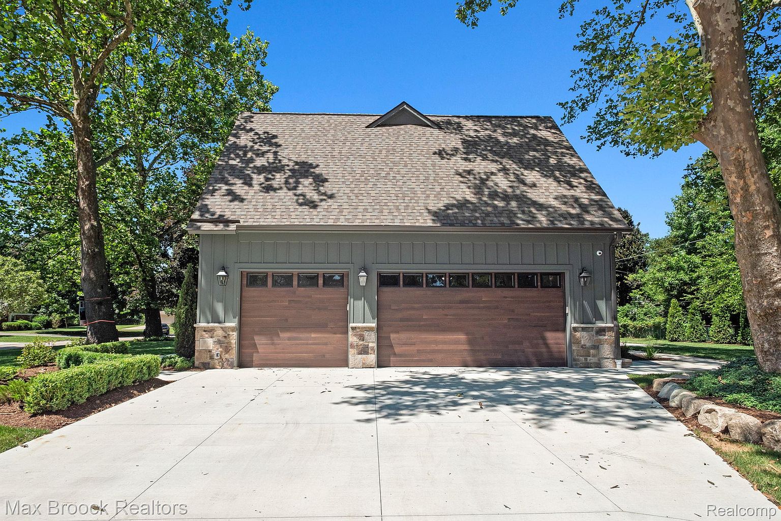 This image showcases a detached two-car garage with a stylish design. The garage features wooden doors, stone accents, and exterior lighting fixtures. The driveway is concrete and well-maintained, and the surrounding landscaping includes mature trees and manicured shrubs, enhancing the property's curb appeal.