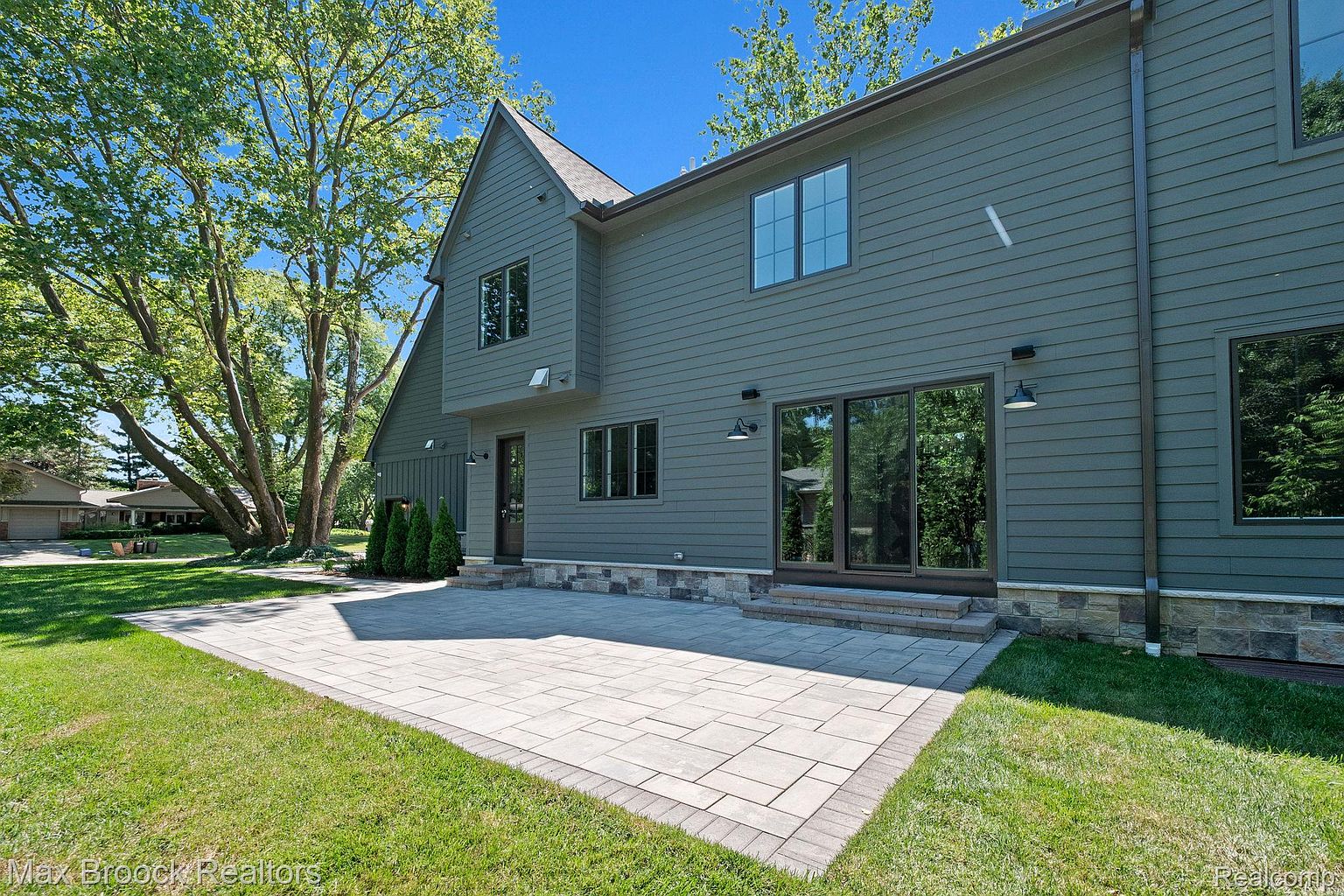 This image showcases the rear exterior of a two-story home with a modern design. The house features gray siding, dark-framed windows, and a stone patio that extends into a well-maintained lawn. Mature trees provide shade and enhance the property's appeal, creating a serene and private backyard space.