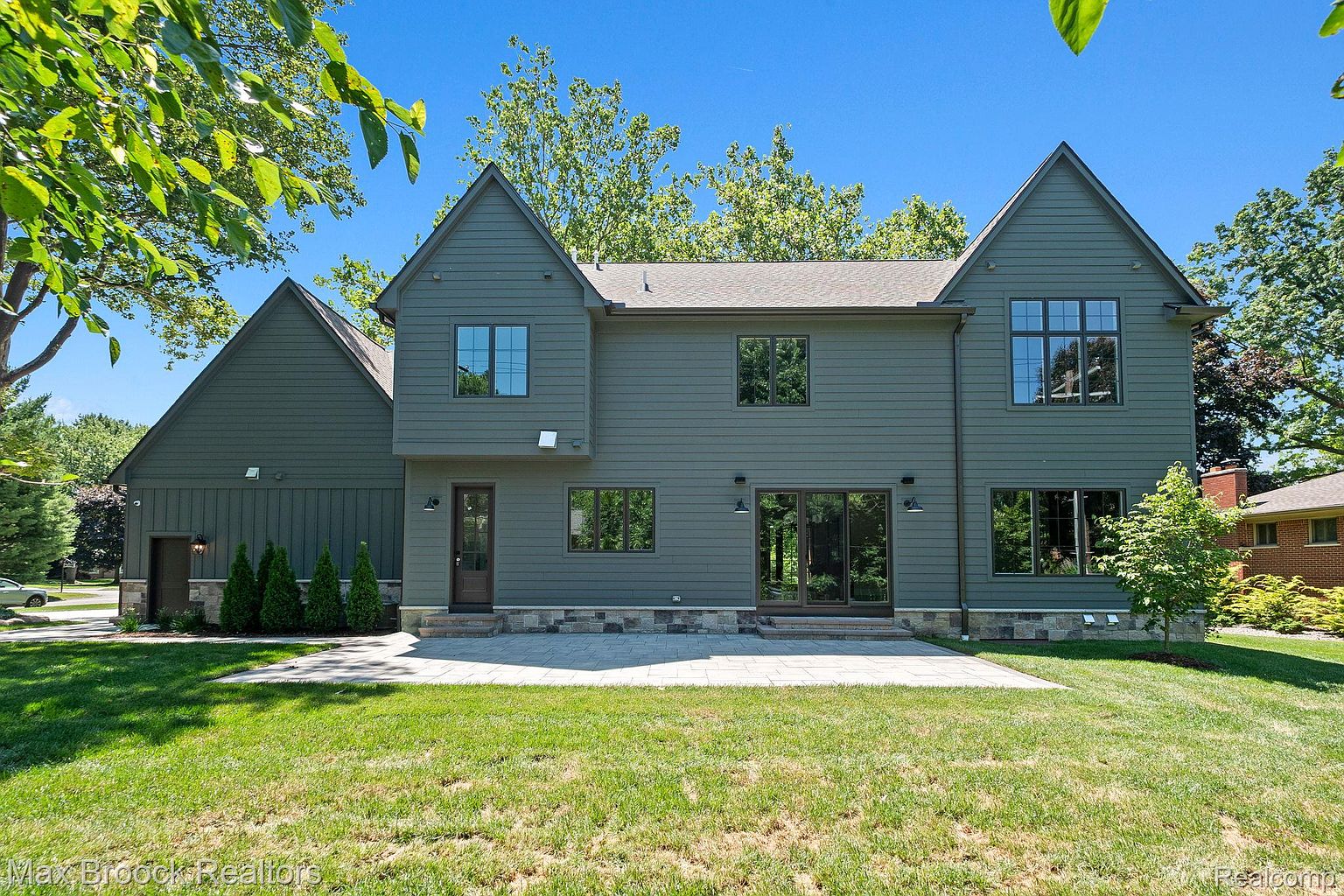 This is a rear view of a two-story house with a dark green exterior and a gray roof. The house features multiple windows and sliding glass doors that lead to a stone patio and a well-maintained lawn. The architecture includes gabled rooflines and a combination of siding and stone accents, creating a sophisticated and modern aesthetic.
