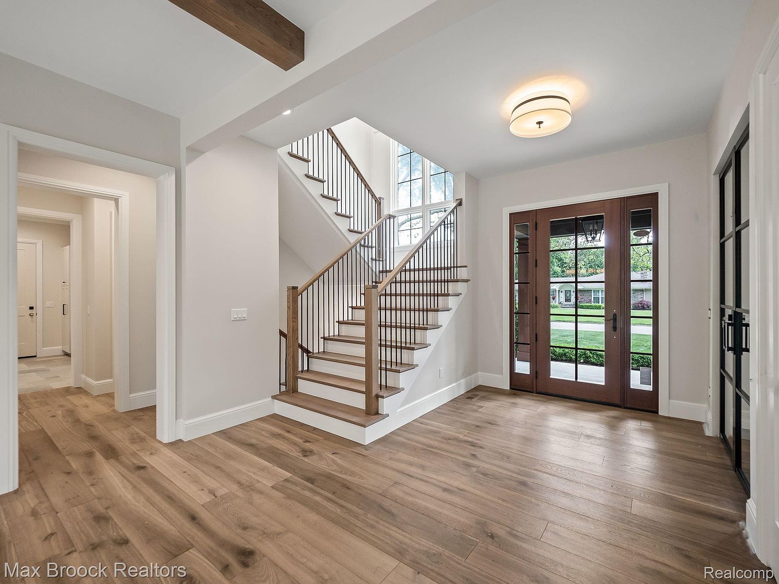 This interior shot showcases a bright and spacious entryway featuring hardwood flooring, a staircase with dark iron railings and light wood treads, and a set of double doors leading to the outside. Natural light floods the area through the doors and a window above the staircase, creating a welcoming and elegant atmosphere. The neutral color palette and clean lines contribute to a modern aesthetic.