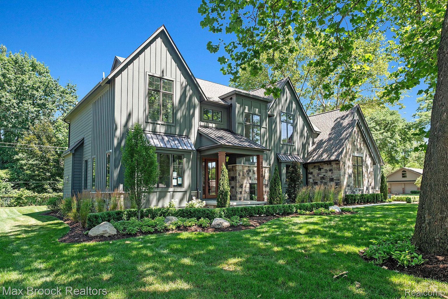 This is a front exterior view of a two-story house with a well-manicured lawn. The house features a combination of gray vertical siding and stone accents, with multiple gabled rooflines and dark-framed windows. The landscaping includes trimmed hedges, ornamental grasses, and mature trees, creating a welcoming and upscale curb appeal.