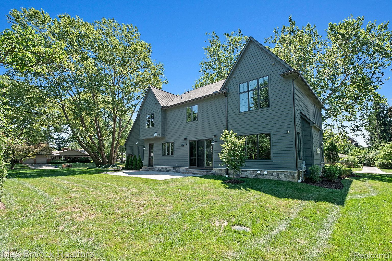 This is a rear view of a modern two-story house with a dark gray exterior. The house features multiple windows, a patio, and a well-maintained lawn. Mature trees surround the property, providing shade and privacy, enhancing the overall appeal of the outdoor space.