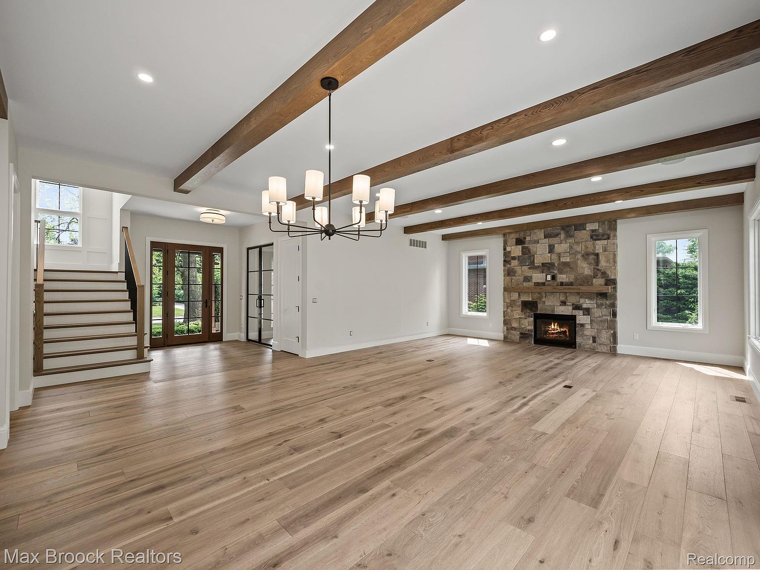 This is a spacious living room featuring hardwood floors, exposed wooden beams, and a stone fireplace. The room is well-lit with natural light from the windows and recessed lighting. A modern chandelier hangs in the center of the room, adding a touch of elegance.