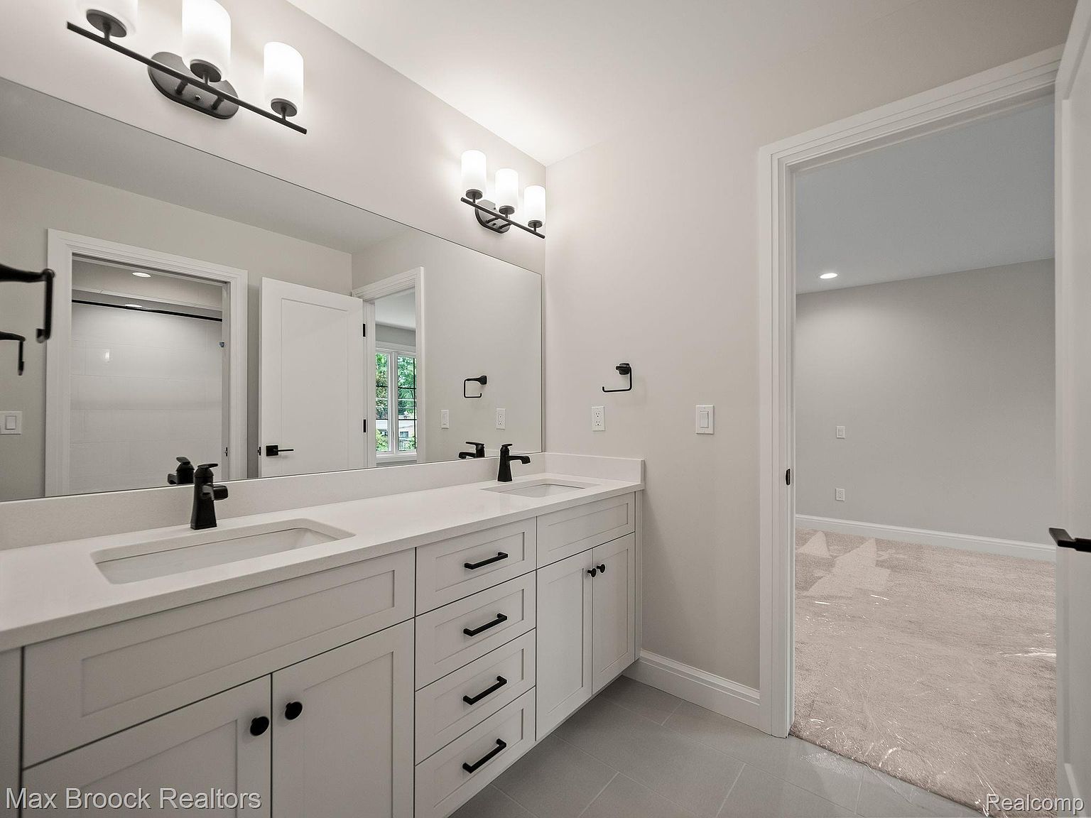 This is a well-lit primary bathroom featuring a double vanity with white countertops and dark hardware. A large mirror spans the length of the vanity, reflecting the lighting fixtures above. The bathroom has a clean, modern aesthetic with gray flooring and a doorway leading to another room.