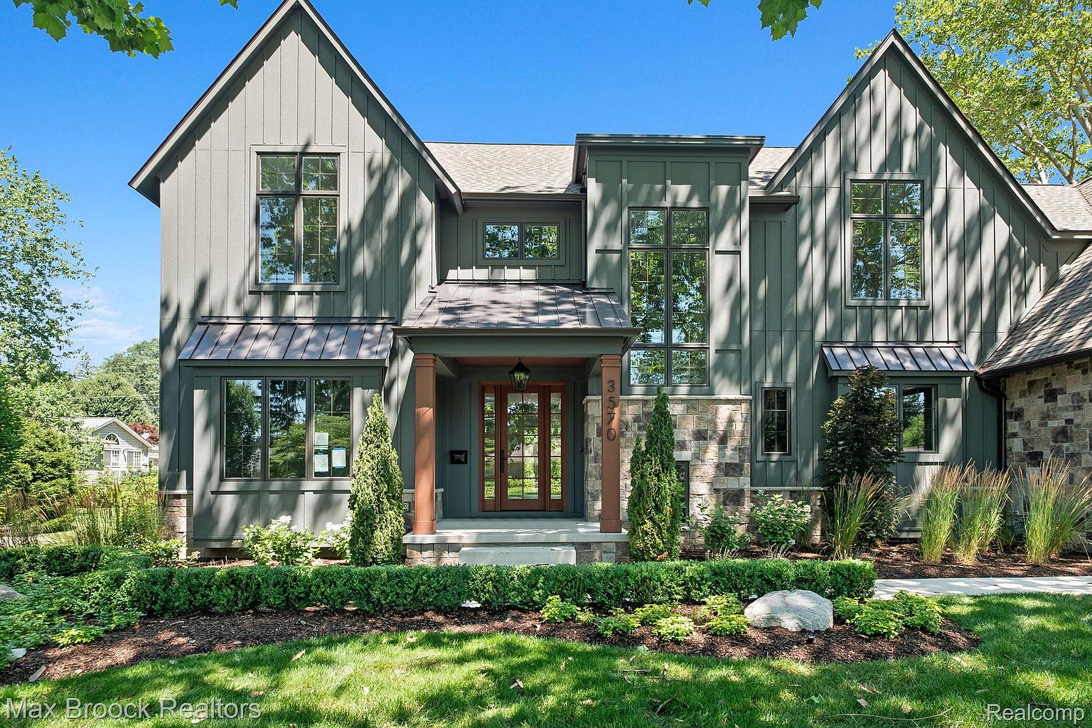 This is a front exterior view of a modern home with a striking facade. The house features a combination of vertical siding and stone accents, creating a visually appealing texture. Large windows provide ample natural light, and the well-manicured landscaping enhances the curb appeal, making it an inviting property.