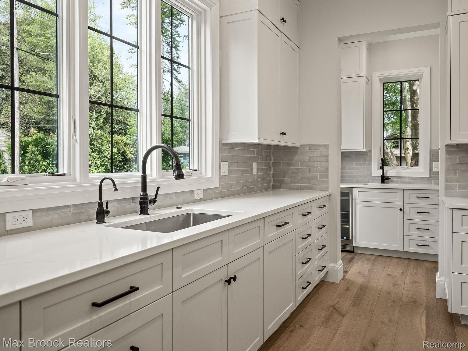 This is a bright and modern kitchen featuring white cabinetry, light gray backsplash, and stainless steel appliances. The kitchen has a large window overlooking a green outdoor space, providing natural light. The countertops are a clean white, complementing the overall minimalist design.