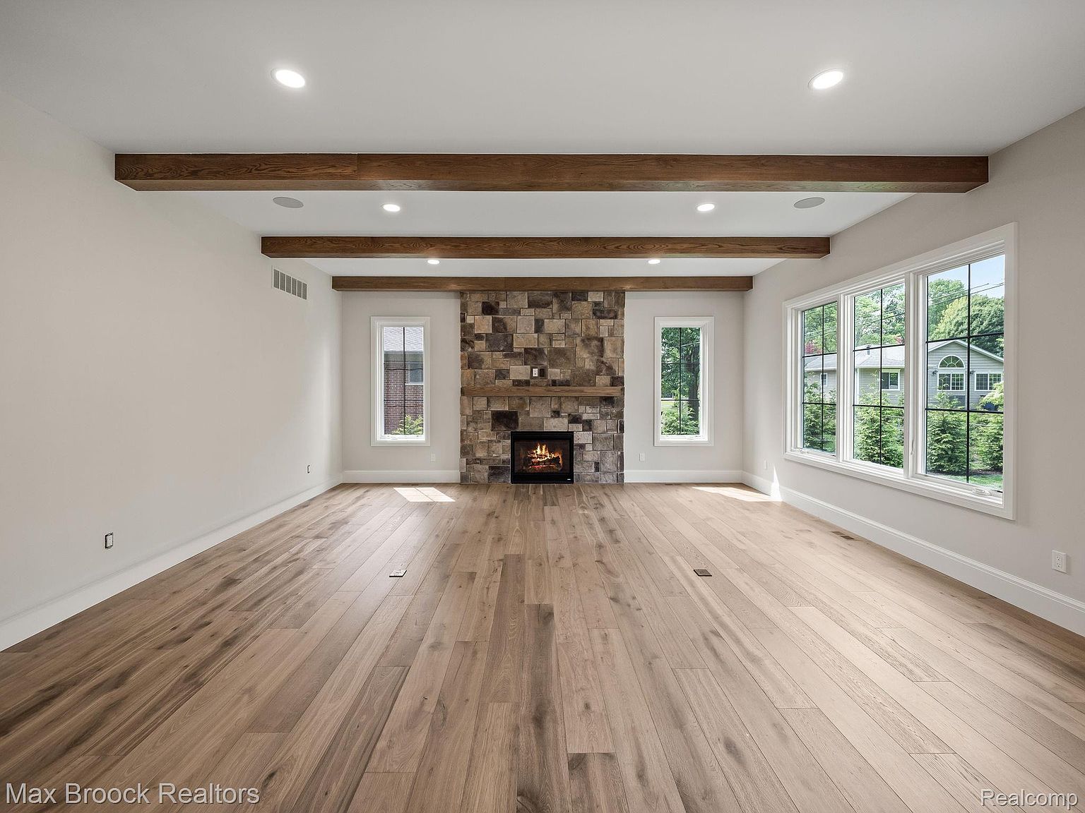This is a spacious living room featuring hardwood floors and a stone fireplace. The room has large windows that provide ample natural light and wooden beams on the ceiling add a rustic touch. The overall impression is clean, modern, and inviting.