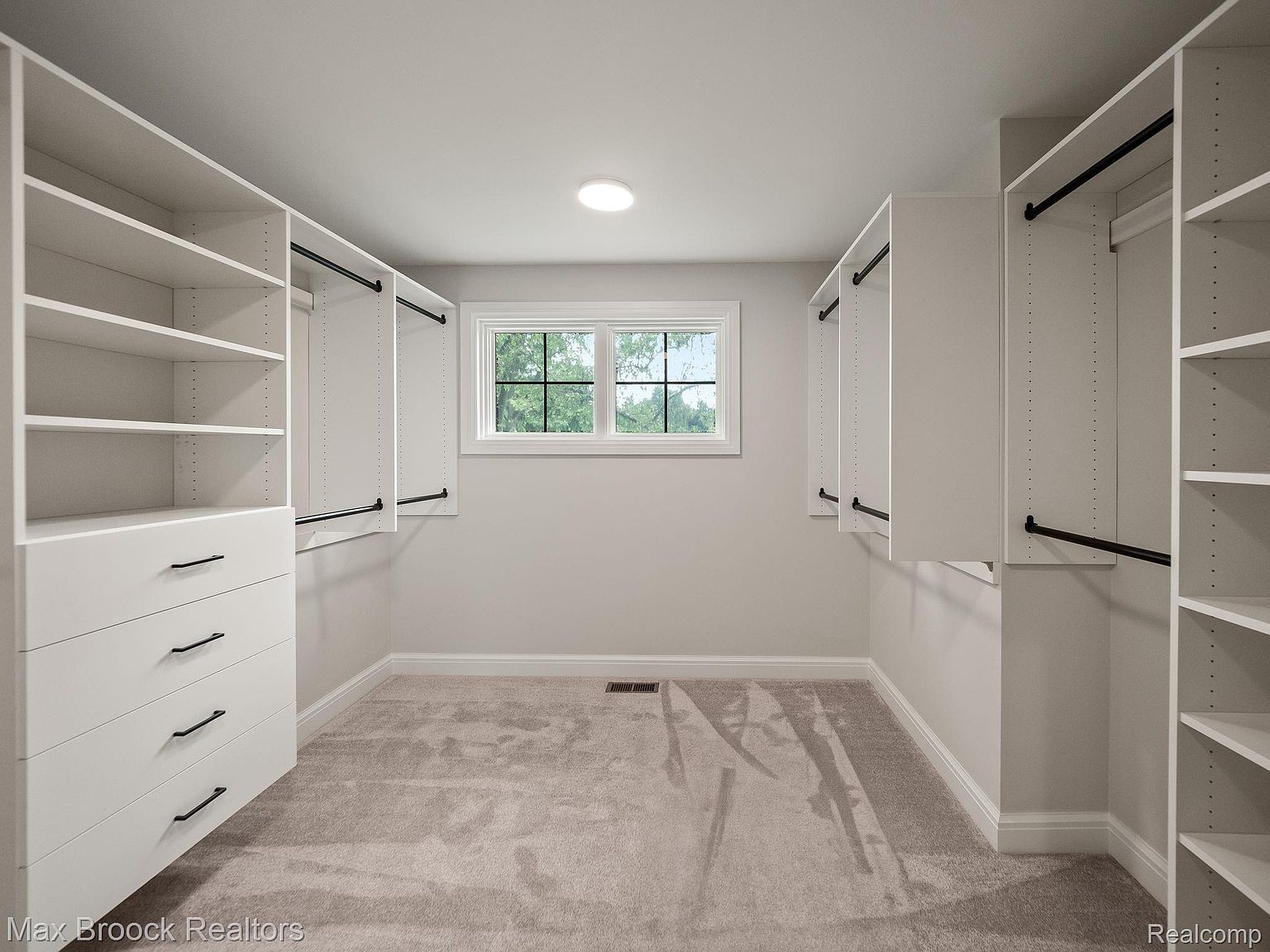 This is a well-organized walk-in closet featuring custom shelving and drawers in a neutral color palette. The closet includes hanging rods and ample storage space, illuminated by a central ceiling light and natural light from a window. The overall impression is clean, spacious, and designed for efficient storage.