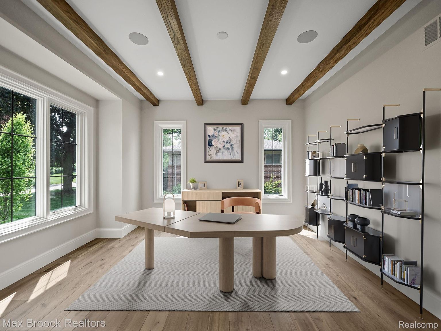 This is an interior shot of a well-lit home office featuring a unique, light-colored wooden desk with cylindrical supports, set upon a large gray rug. The room is accented with exposed wooden beams on the ceiling and a modern black shelving unit against the right wall, providing ample storage and display space. Natural light floods the room through multiple windows, creating a bright and inviting workspace.