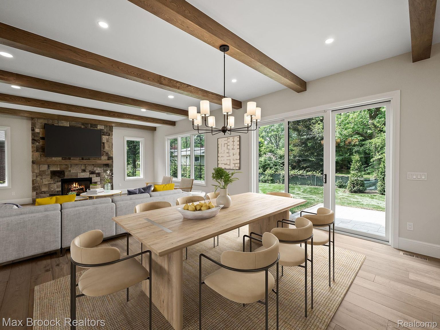 This is an interior shot of a dining room featuring a long wooden table with modern chairs. A contemporary chandelier hangs above the table, and large sliding glass doors provide ample natural light and a view of the backyard. The room is decorated in neutral tones with wooden beams on the ceiling, creating a warm and inviting atmosphere.