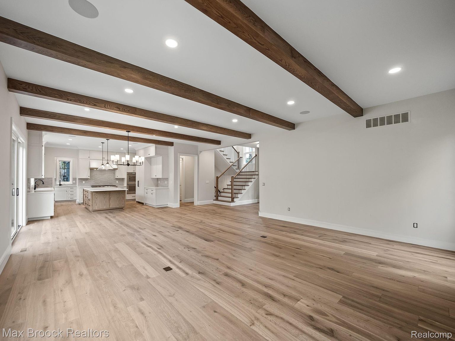 This is a spacious open-concept living area featuring hardwood floors, exposed wooden beams, and recessed lighting. The space seamlessly connects to a modern kitchen with white cabinetry and a central island, as well as a staircase leading to the upper level. The overall impression is one of contemporary elegance and comfortable living.
