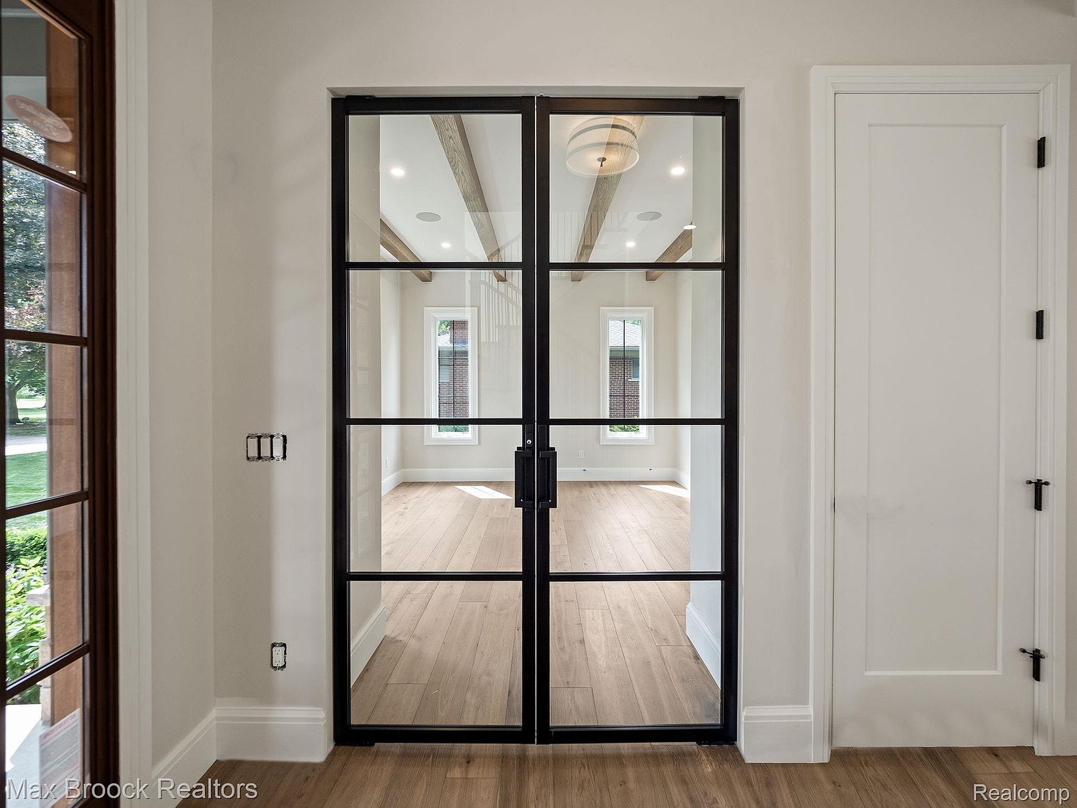 The image showcases an interior hallway featuring modern black-framed glass double doors that lead into another room, allowing natural light to flood the space. A white door is visible to the right, and the hallway is finished with light hardwood flooring. The overall impression is clean, bright, and contemporary, highlighting the home's architectural details.