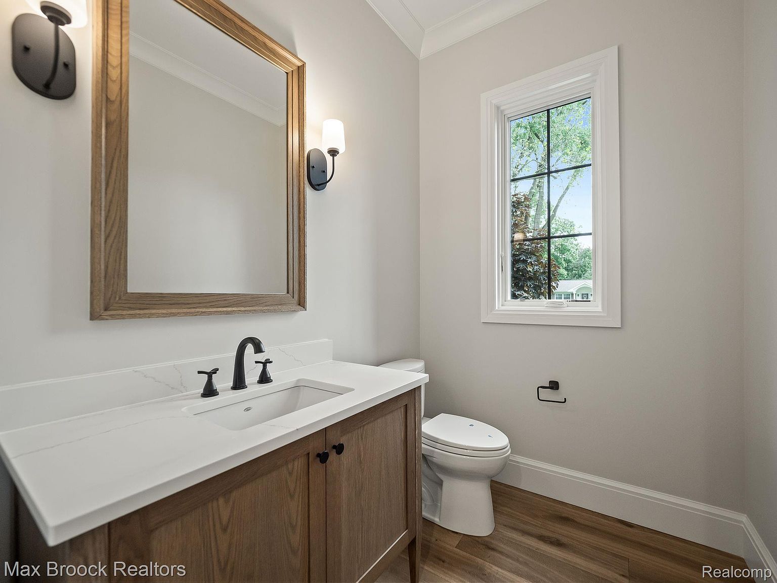 This is a well-lit guest bathroom featuring a wooden vanity with a white countertop and black hardware. A framed mirror hangs above the sink, flanked by sconce lighting. A toilet sits next to a window that provides natural light, and the flooring appears to be wood-look tile, creating a clean and modern aesthetic.