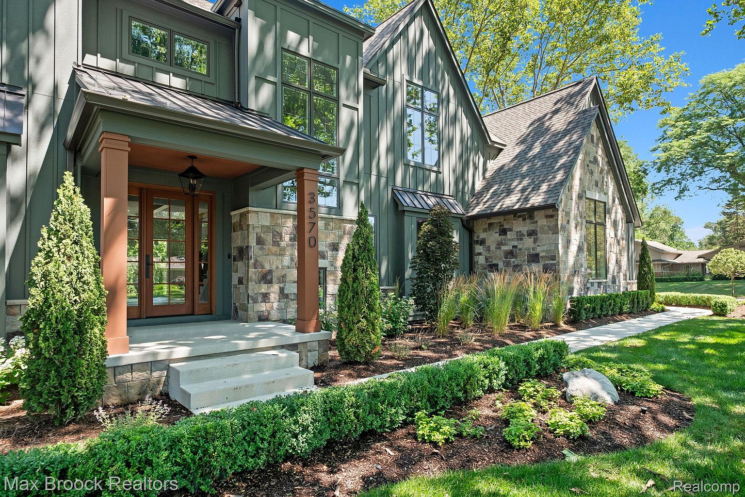 This is a front exterior view of a two-story house with a combination of green siding and stone accents. The entryway features a covered porch with wooden support columns and a double-door entrance. Landscaping includes manicured hedges, ornamental grasses, and small trees, enhancing the curb appeal of the property.