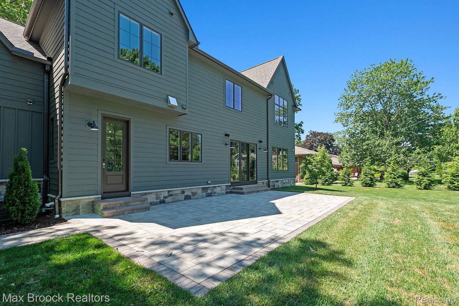 This is a rear exterior view of a two-story house, showcasing a well-maintained backyard. The house features green siding, dark-framed windows, and a stone patio that extends from the back doors. The yard is lush with green grass and mature trees, creating a serene and inviting outdoor space.