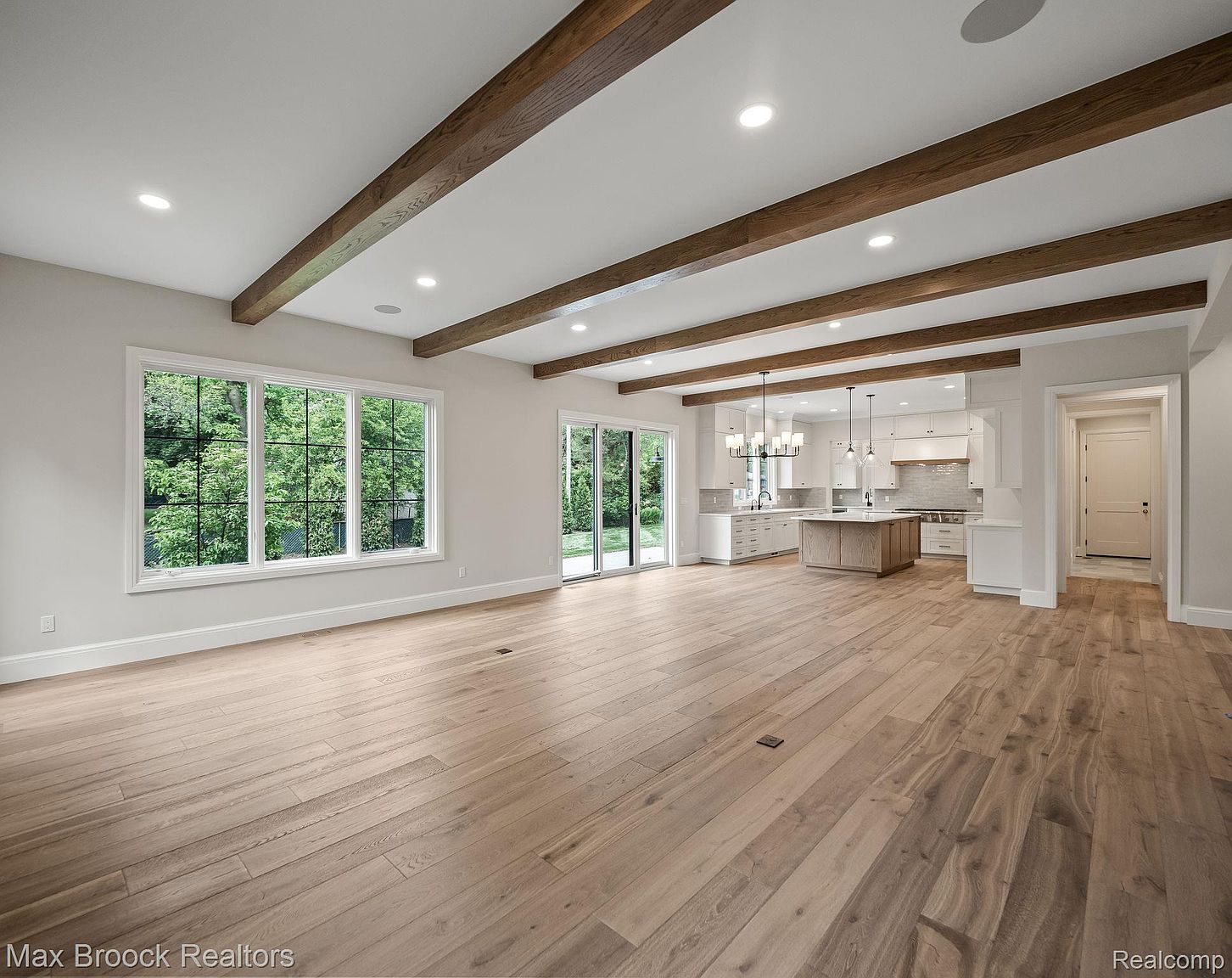 This is an interior shot of a spacious living room that seamlessly transitions into a modern kitchen. The living room features hardwood floors, wooden ceiling beams, and a large window offering natural light. The kitchen boasts white cabinetry, a central island, and pendant lighting, creating an open and inviting atmosphere.