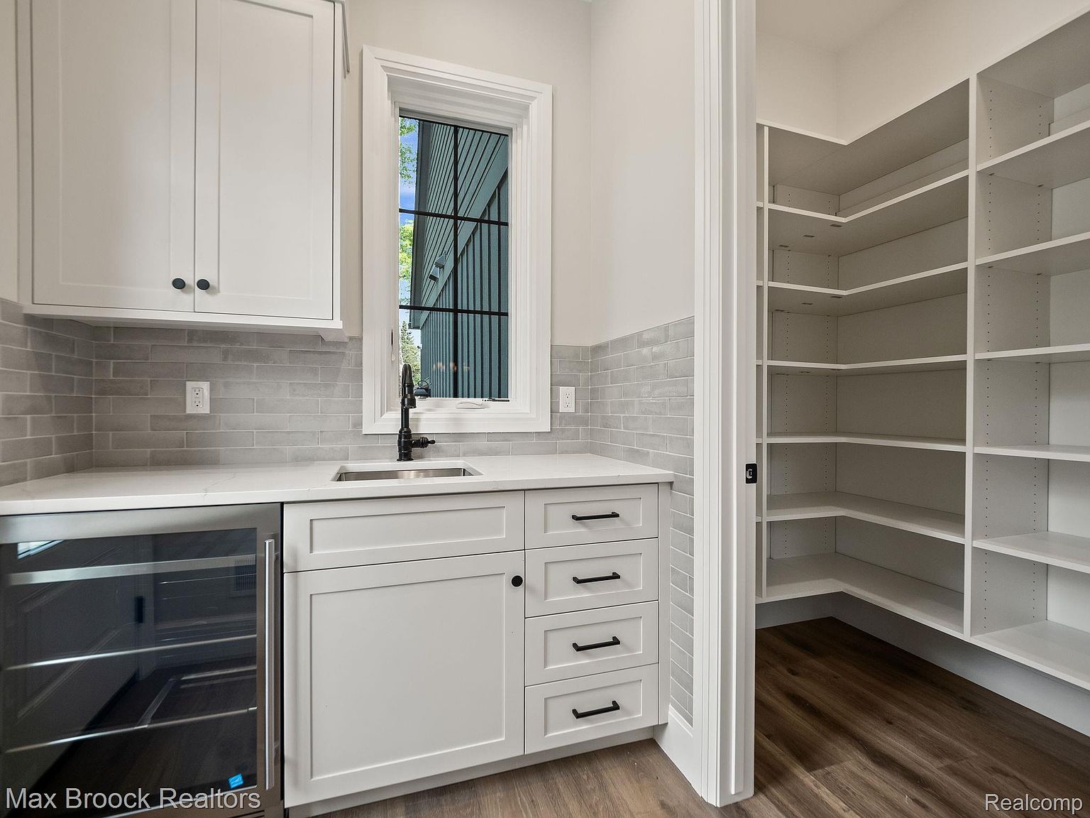 This interior shot showcases a well-organized pantry with white shelving and drawers, complemented by a gray backsplash and light countertops. A small sink and beverage cooler are integrated into the design, enhancing functionality. The pantry is adjacent to a storage area with open shelving, suggesting ample space for food and household items.