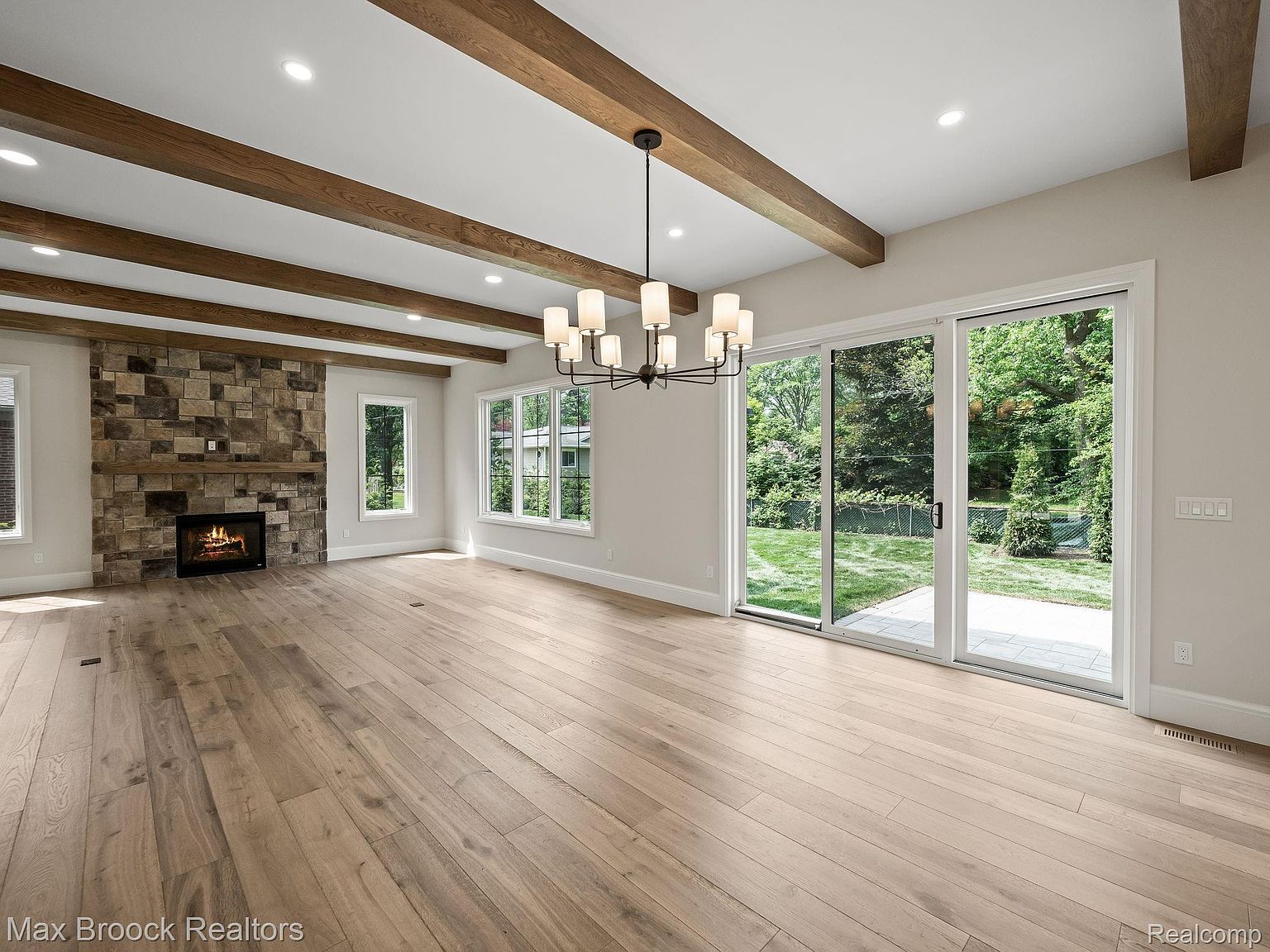 This is a bright and spacious living room featuring hardwood floors, a stone fireplace, and exposed wooden beams on the ceiling. Large sliding glass doors provide ample natural light and access to the outdoors. A modern chandelier hangs in the center of the room, adding a touch of elegance.