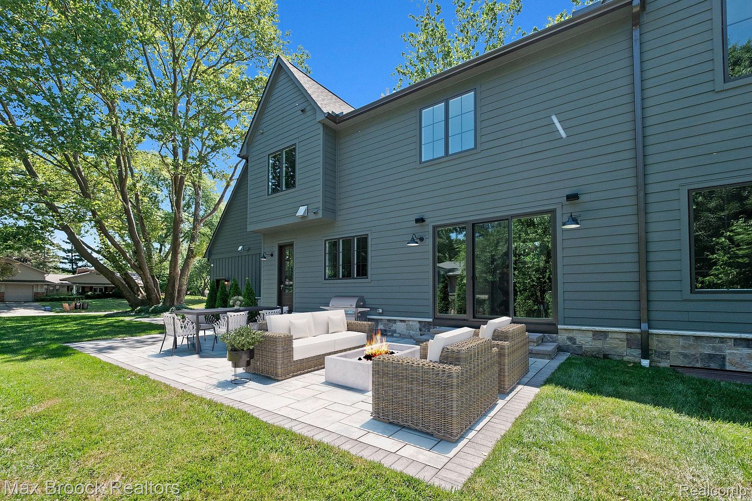 This image showcases a beautifully designed outdoor patio area, featuring comfortable wicker seating arranged around a modern fire pit. The patio is constructed with light-colored pavers and bordered by lush green grass, creating an inviting space for relaxation and entertainment. The house's exterior, with its gray siding and dark-framed windows, provides a stylish backdrop to the outdoor living area.