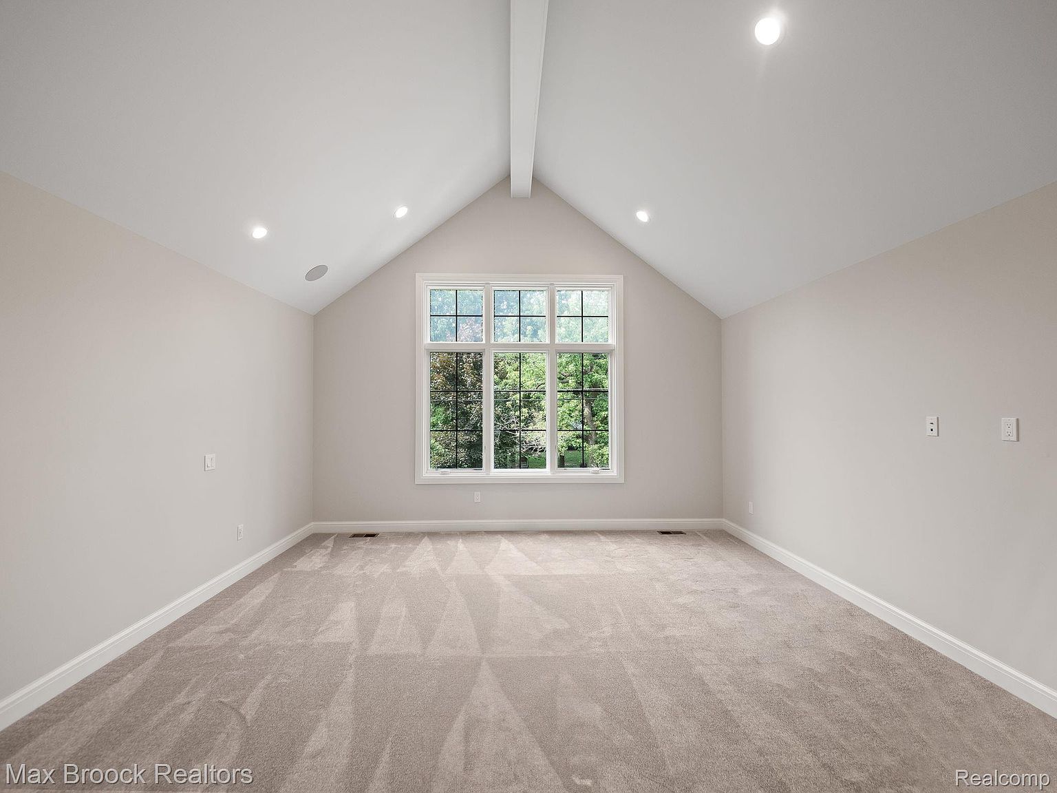 This is an interior shot of a spacious bedroom featuring neutral-toned walls and carpet. A large window provides ample natural light, highlighting the room's clean and airy atmosphere. The vaulted ceiling with a central beam adds architectural interest, making it an inviting space.