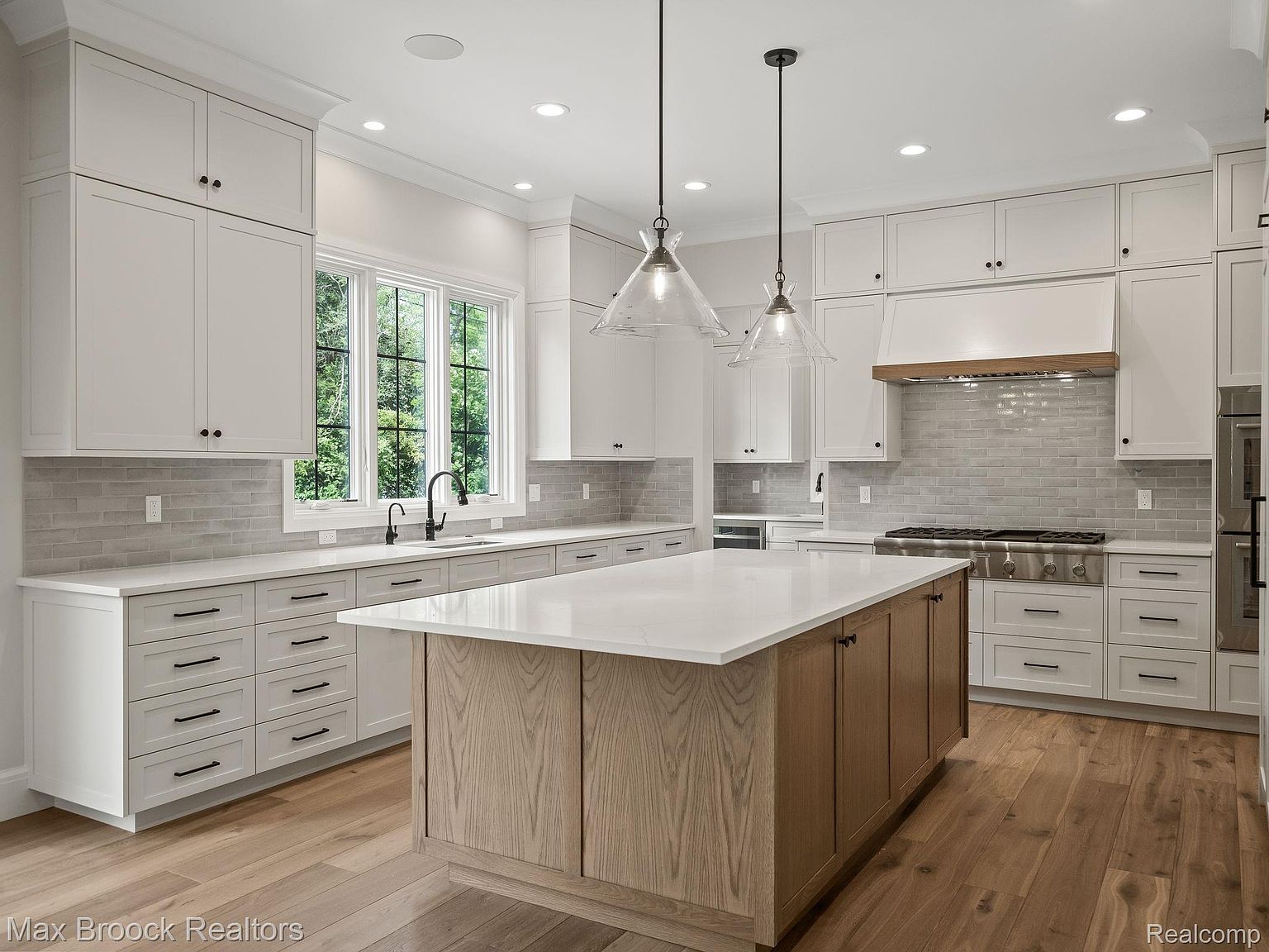 This is a bright and modern kitchen featuring white cabinetry, a large center island with a light wood base and white countertop, and stainless steel appliances. The kitchen is well-lit with recessed lighting and pendant lights over the island, and a large window provides natural light and a view of the outdoors. The hardwood flooring adds warmth to the space, creating an inviting atmosphere.