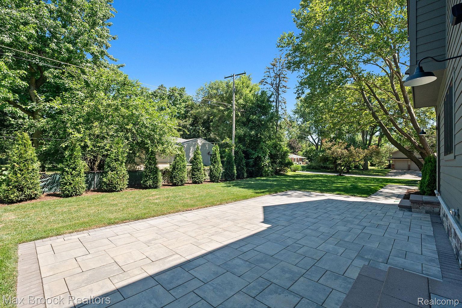 This image showcases a well-maintained patio area, featuring a paved surface that extends from the side of a house into a lush green lawn. The patio is bordered by manicured shrubs and trees, creating a private and inviting outdoor space. The clear blue sky enhances the overall appeal, suggesting a perfect setting for relaxation and entertainment.