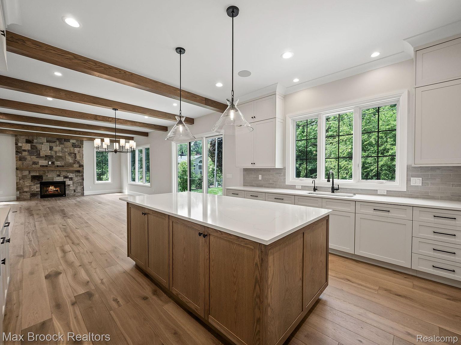 This is a bright and spacious kitchen featuring a large center island with a white countertop and wooden base. The kitchen also includes white cabinetry, stainless steel appliances, and a window overlooking the backyard. The open floor plan extends into a living area with a stone fireplace and wooden beams, creating a warm and inviting atmosphere.