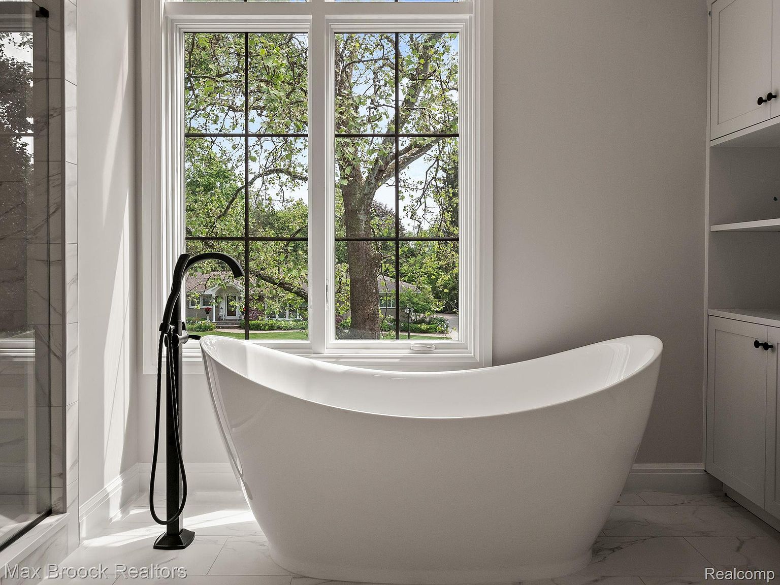 This is a bright and airy primary bathroom featuring a freestanding white bathtub positioned in front of a large window with a view of lush greenery. The room is decorated in neutral tones, with white marble flooring and cabinetry, and a modern black faucet adds a touch of contrast. The overall impression is one of luxury and relaxation.