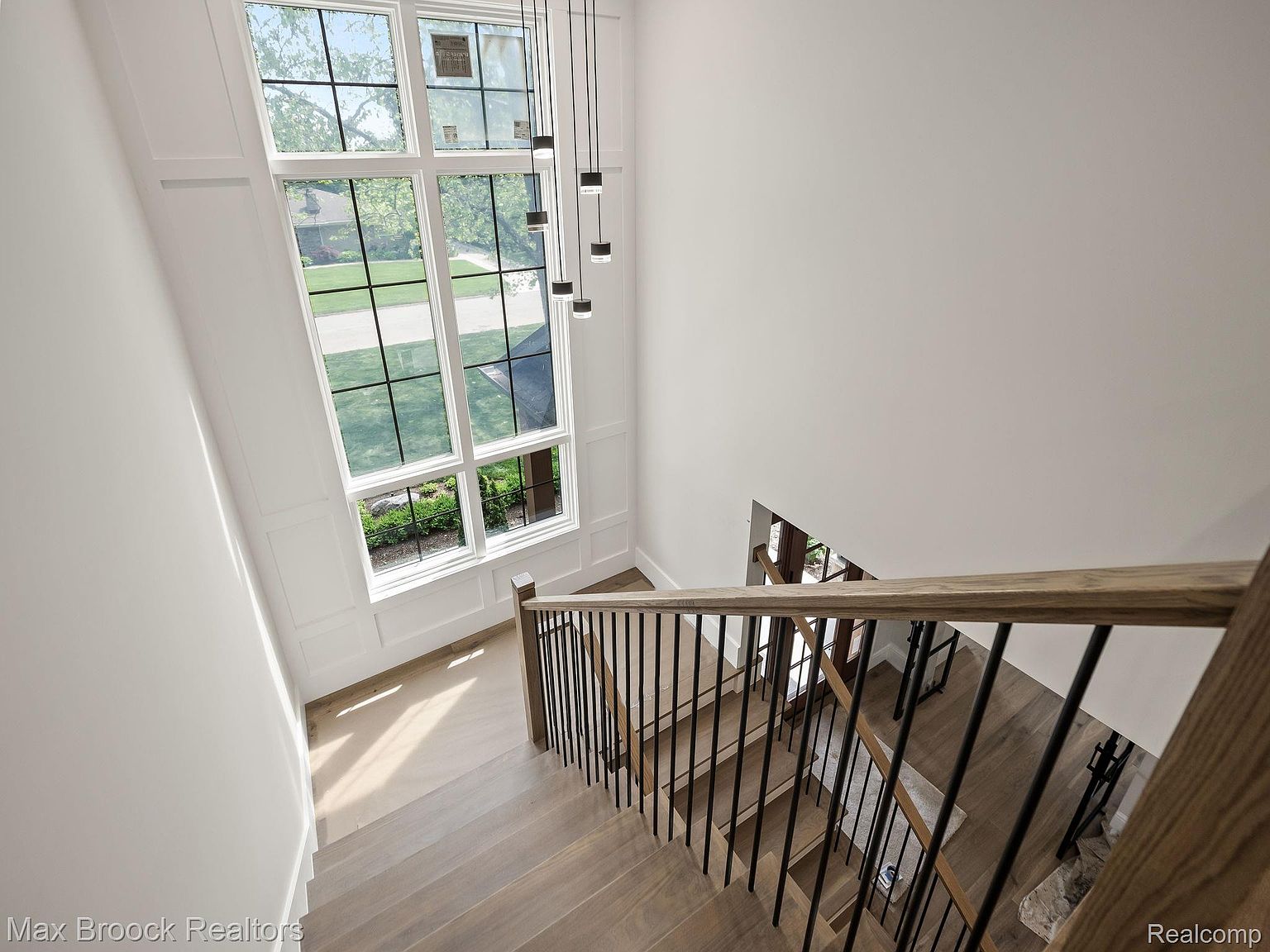 This interior shot showcases a modern staircase with wooden steps and black metal railings, ascending towards a large window that floods the space with natural light. The walls are painted in a neutral tone, enhancing the bright and airy feel of the hallway. The view is from above, looking down the staircase.