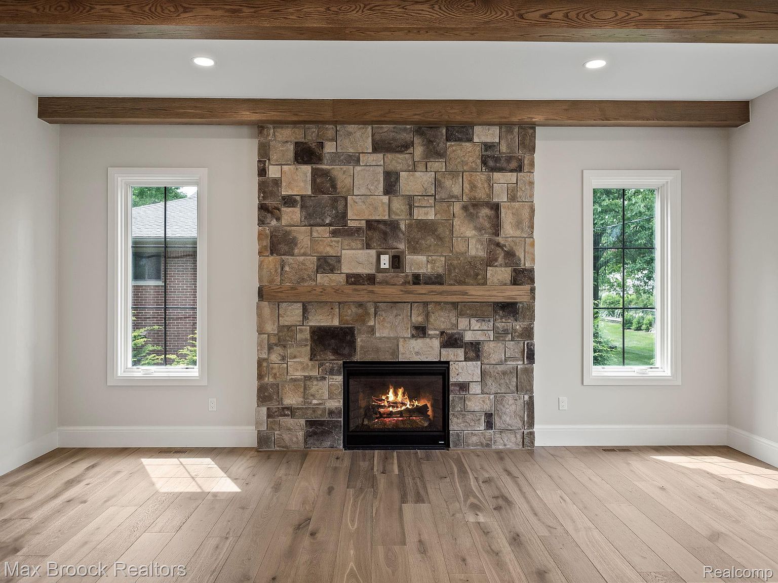 This is an interior shot of a living room, showcasing a stone fireplace as the focal point, flanked by two windows that provide natural light. The room features hardwood flooring and wooden beams on the ceiling, creating a warm and inviting atmosphere. The style is modern rustic, blending natural materials with clean lines.