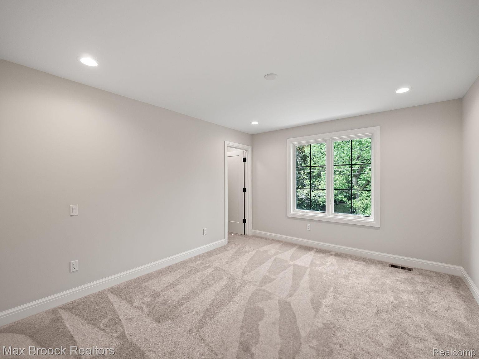 The image showcases a bright and airy bedroom with neutral tones. The room features a large window providing ample natural light, complemented by recessed lighting. The carpet is a light beige with a subtle pattern, and the walls are painted in a soft, warm gray, creating a calming and inviting atmosphere.