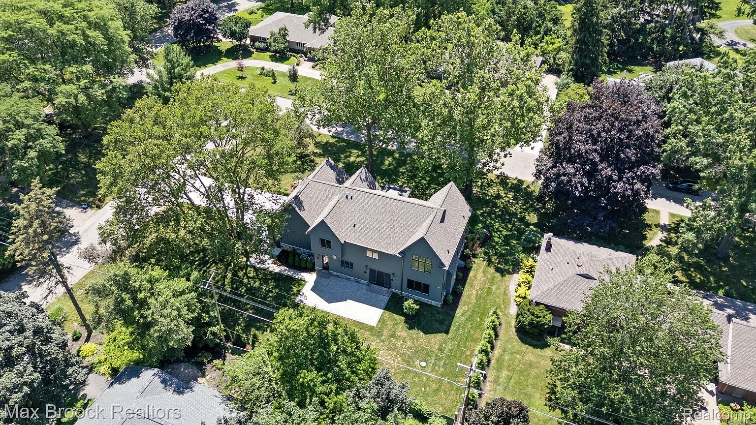 This aerial shot showcases a modern, dark-colored house with a gray roof, nestled among lush green trees. The property features a well-maintained lawn and a concrete patio area. The surrounding landscape includes mature trees and neighboring houses, creating a serene and private setting.
