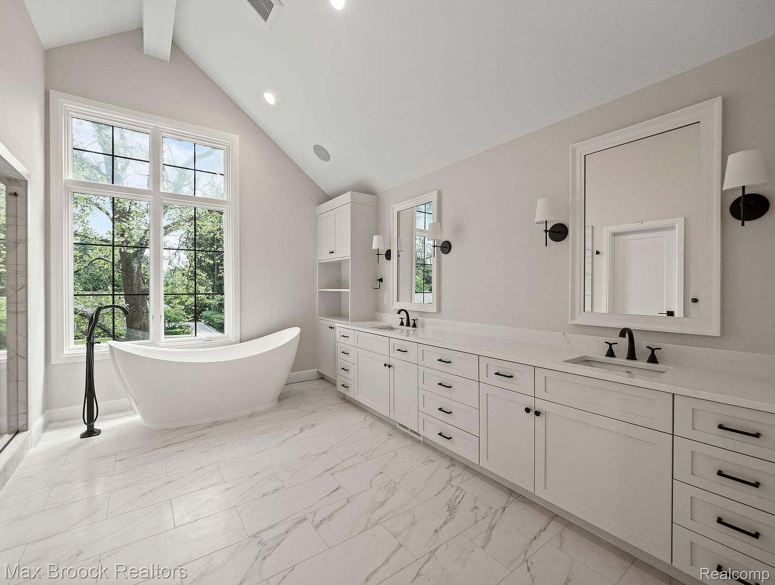 This is a bright and spacious primary bathroom featuring a freestanding white bathtub positioned in front of a large window offering a view of the outdoors. The bathroom also includes a long vanity with dual sinks, white cabinetry with black hardware, and marble-like tile flooring, creating a luxurious and serene atmosphere.